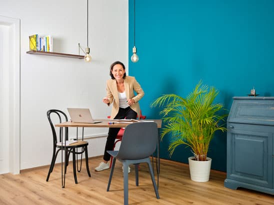 Andrea Elber hinter einem Schreibtisch mit Laptop stehend, drumherum 2 Stühle, eine Palme, ein Sideboard